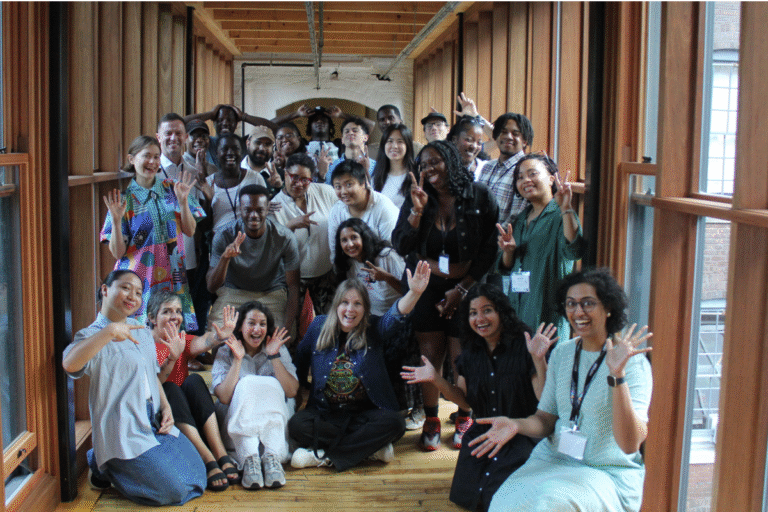A large group photo of participants standing and sitting together in a covered wooden walkway, smiling and raising their hands in celebratory poses.