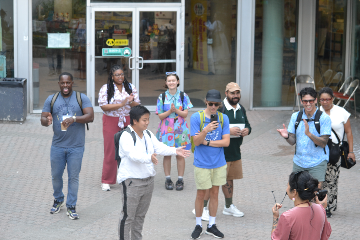A group of people standing in an outdoor plaza area in front of glass doors. Some are holding phones or drinks, and they appear to be observing or participating in an activity.