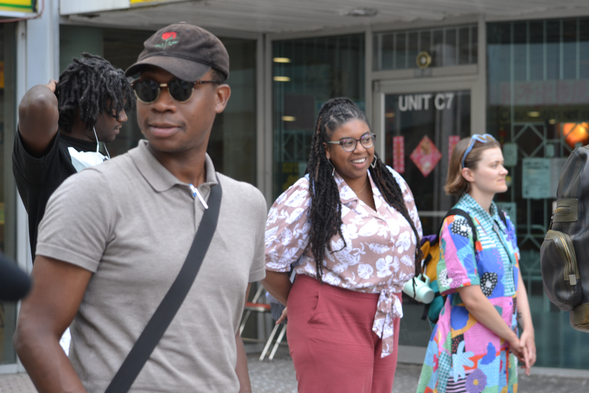 People standing outside a storefront with glass windows, wearing casual clothing and backpacks. They appear to be listening or waiting as they face toward the camera.