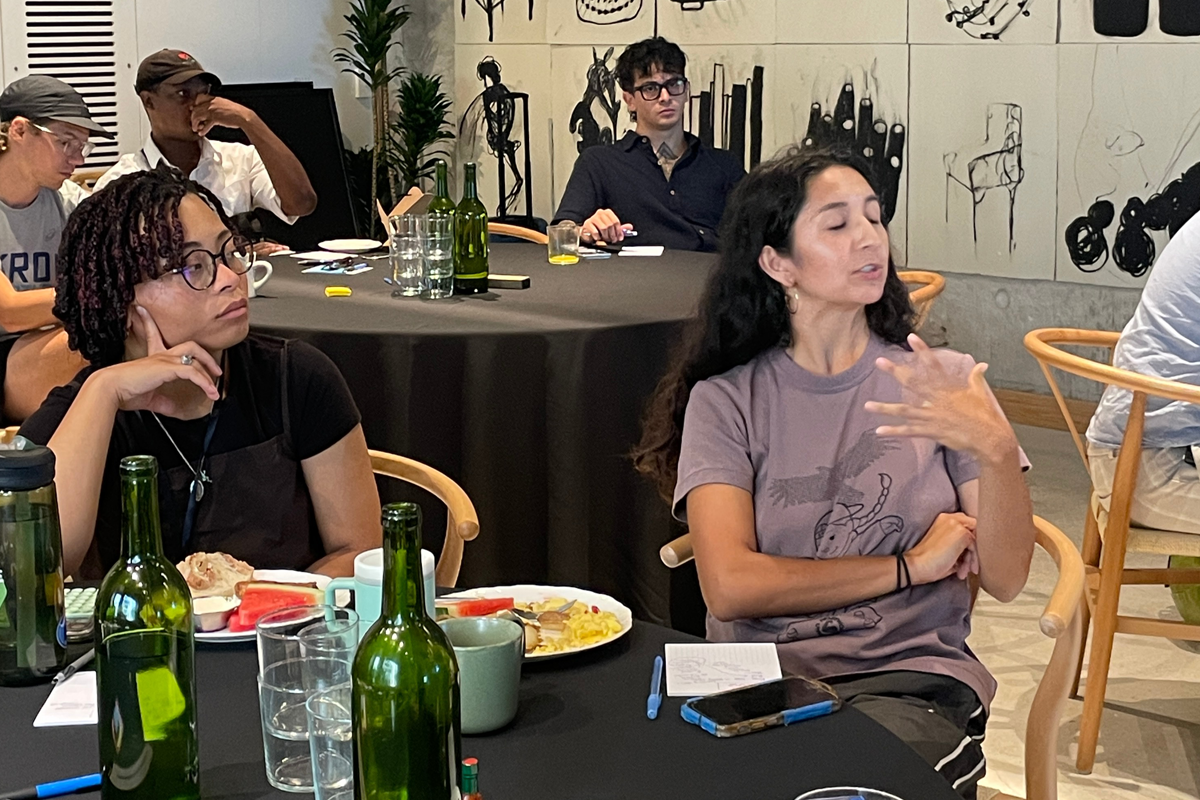 A small group of people seated around circular tables during an indoor session. Notebooks, plates of food, and water bottles are on the tables while attendees listen and talk.